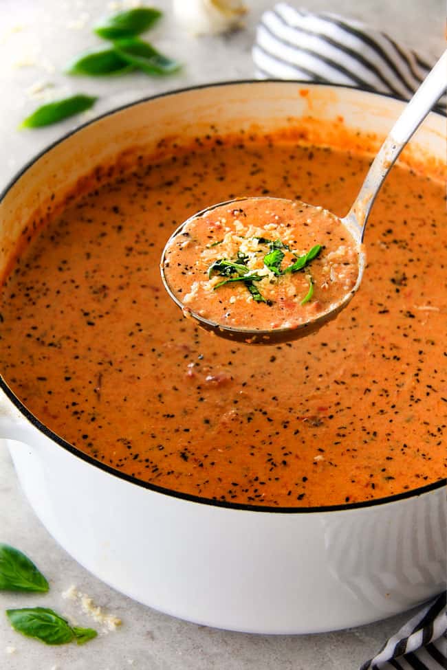 This Tomato Basil Soup recipe is our family favorite! Its super easy without any chopping! bursting with flavor and I love the addition of Parmesan! a ladle of Creamy Tomato Basil Soup raised above a big soup pot.