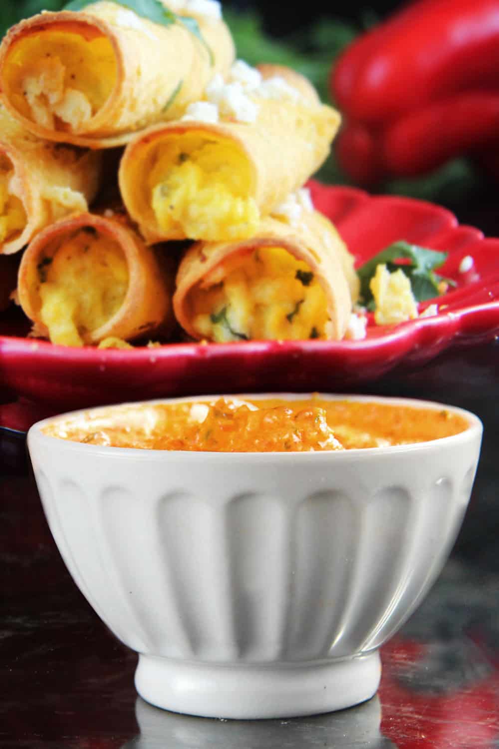 Breakfast Taquitos on a red plate focusing on the red pepper and feta dipping sauce in a white serving bowl.