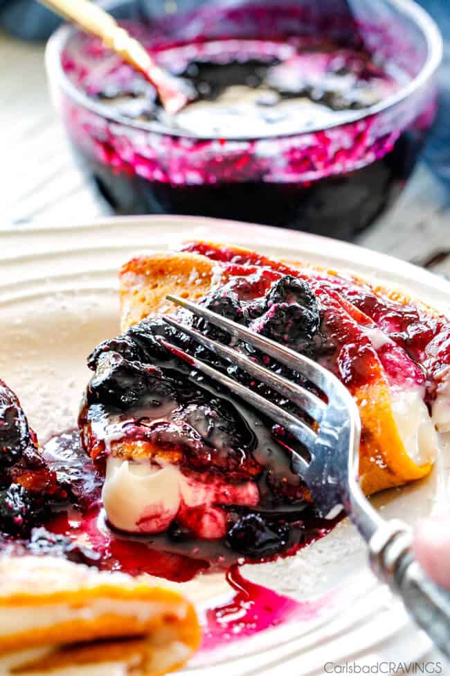 A fork cutting into a piece of Carrot Cake Crepes with Cream Cheese Filling and Blueberry Sauce.