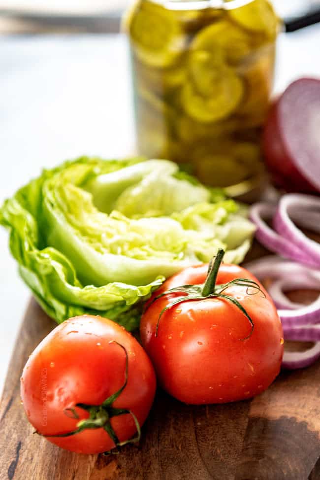 showing how to make burgers by lining up condiments of tomatoes, lettuce, red onions and pickles on a cutting board