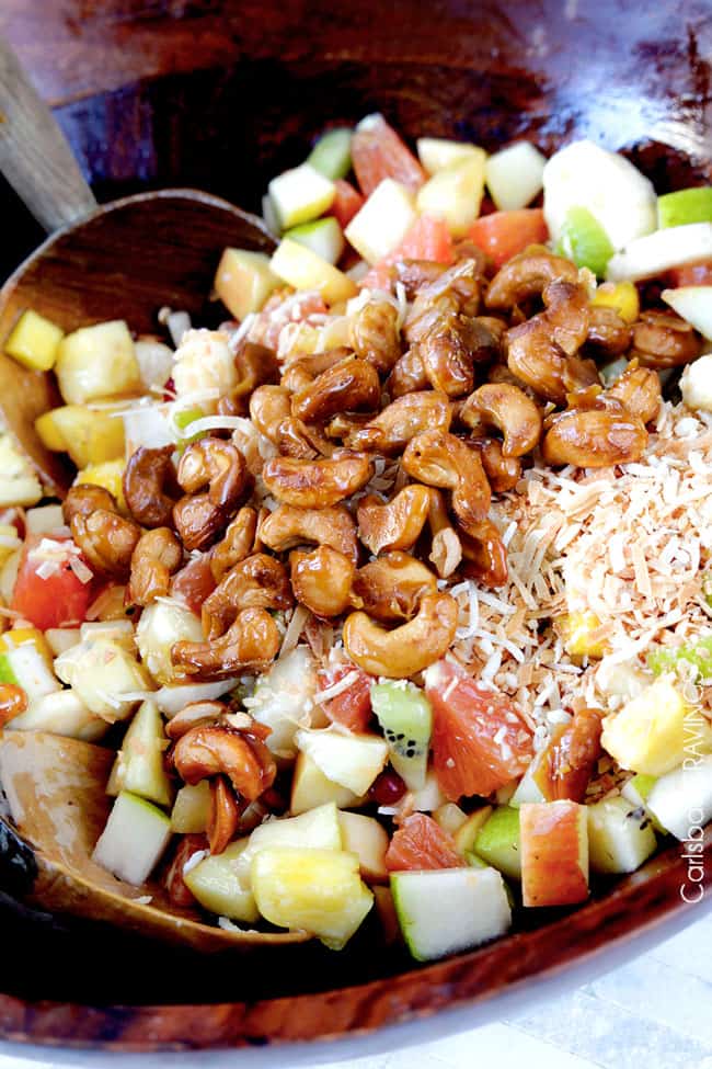 showing how to make tropical fruit salad by adding coconut and cashews to salad before tossing