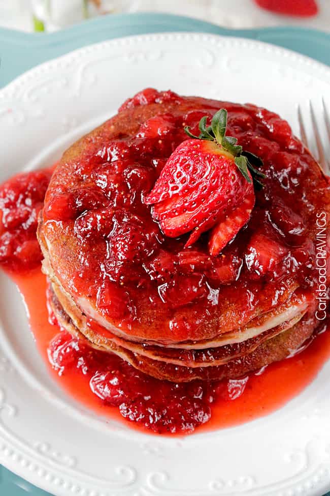 Stack of pancakes doused in strawberry topping on a white plate.