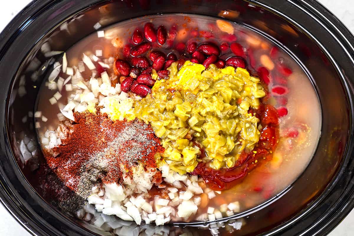 showing how to make chicken chili by adding chicken, beans, onions, garlic, seasonings and green chilies to crockpot