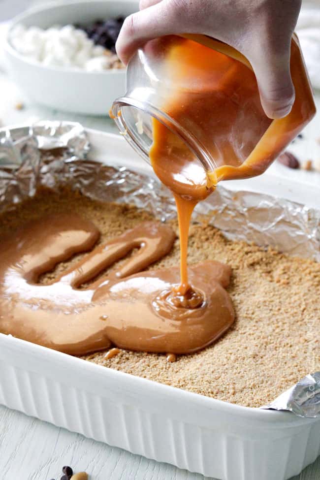 Showing how to make Caramel Nut S'mores Bars by pouring caramel sauce over the graham base.