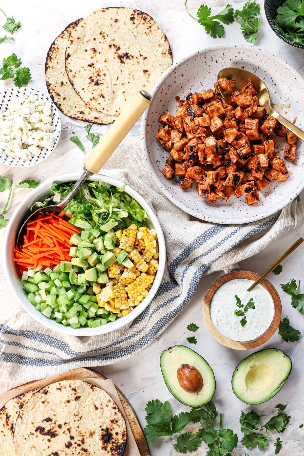 top view of buffalo chicken taco ingredients: buffalo chicken, tortillas, avocado and corn and carrot and celery topping, and blue cheese cilantro ranch