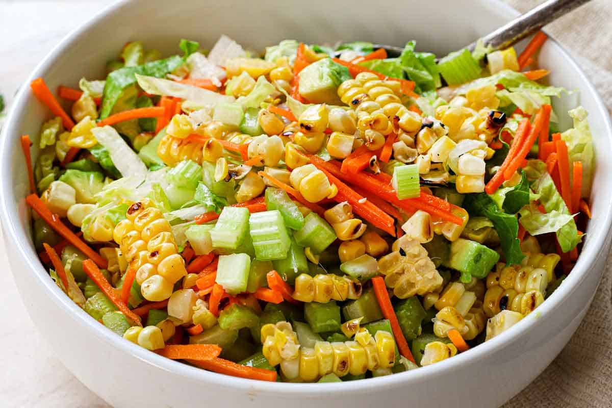 showing how to make buffalo chicken tacos by tossing the corn, lettuce, carrots, celery and avocado together in a bowl