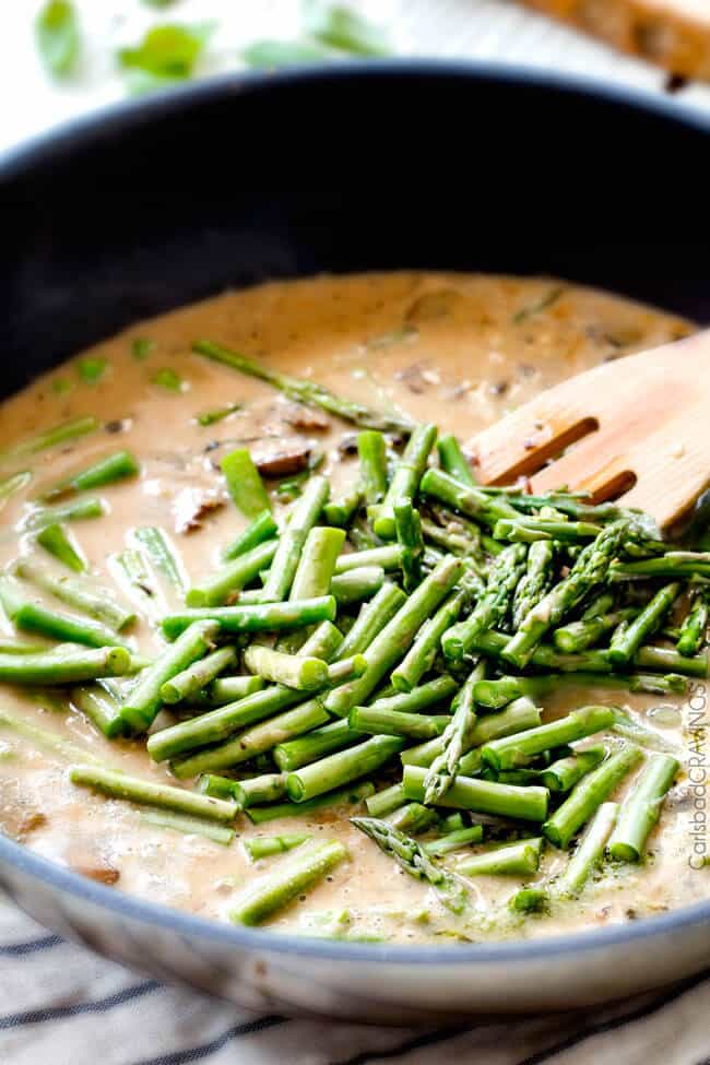 showing how to make pesto chicken by adding asparagus to the skillet.