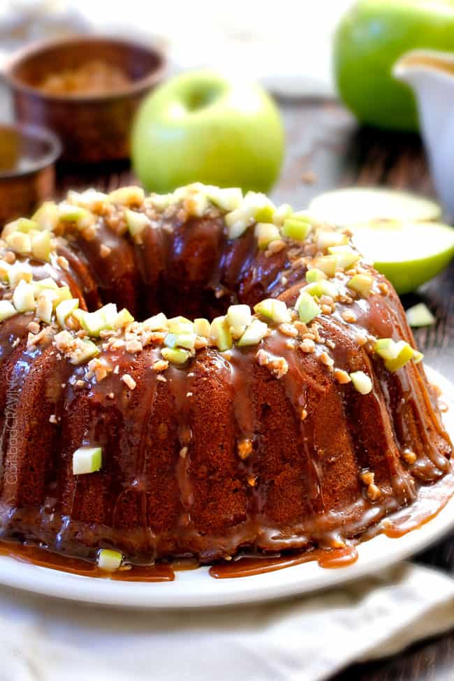 apple bundt cake on a white plate. The cake is drizzled with caramel sauce and topped with toffee bits and chopped apple.