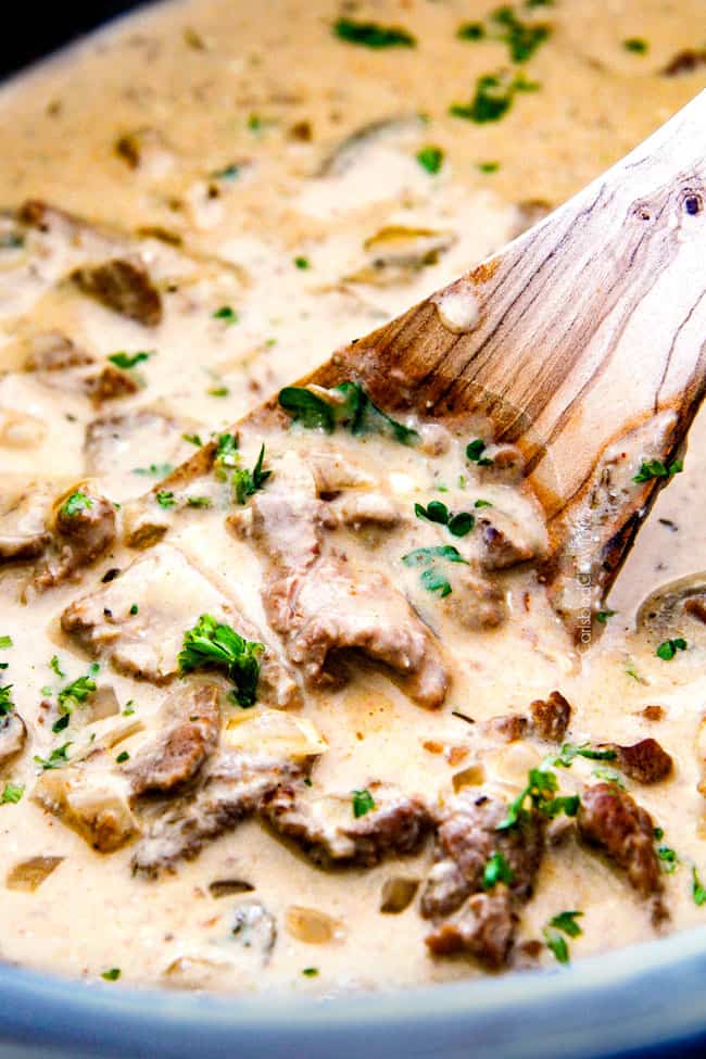This is by far my family's favorite Slow Cooker Beef Stroganoff recipe! An ultra rich and creamy, amazingly flavorful sauce (without any "cream of" anything!), crazy tender meat all made in the crockpot for a gourmet meal with hardly any effort! I love serving this for holidays like Christmas! up close of crock pot Beef Stroganoff in the crock pot with a wooden spoon in rich gravy garnished with parsley