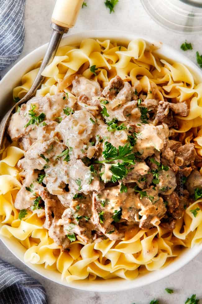 This is by far my family's favorite Slow Cooker Beef Stroganoff recipe! An ultra rich and creamy, amazingly flavorful sauce (without any "cream of" anything!), crazy tender meat all made in the crockpot for a gourmet meal with hardly any effort! I love serving this for holidays like Christmas! up close top view of slow cooker beef stroganoff in a white bowl with a silver servings spoon