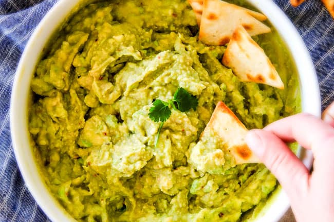 This creamy Avocado Hummus is SO addicting and only 3 ingredients! Its crazy flavorful, healthy and super easy! top view of a hand dipping a chip in healthy avocado hummus