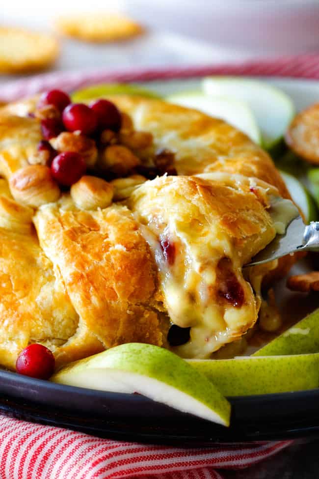 Rich and creamy Baked Brie in Puff Pastry is always a total luxuriously tasting crowd pleaser with minimal effort! (step by step pictures included) It can be prepped in advance then baked when you're ready for company! a serving spoon taking a slice of baked brie in puff pastry with cranberries and jam