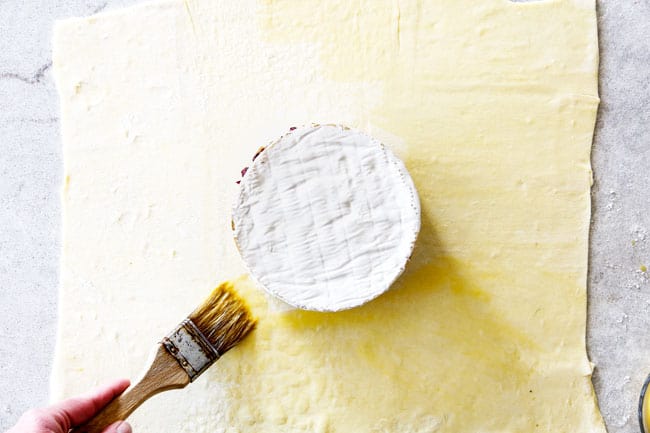 Rich and creamy Baked Brie in Puff Pastry is always a total luxuriously tasting crowd pleaser with minimal effort! (step by step pictures included) It can be prepped in advance then baked when you're ready for company! aaaaaaaaa showing how to make baked brie with puff pastry by laying puff pastry on a flat surface with brie wheel in the center