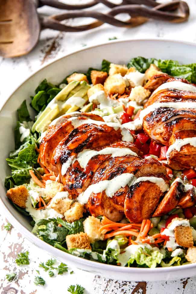 side view of sliced Buffalo Chicken in Buffalo Chicken Salad in a white bowl with blue cheese, celery, carrots, avocado, red bell peppers and lettuce with Blue Cheese Ranch Dressing