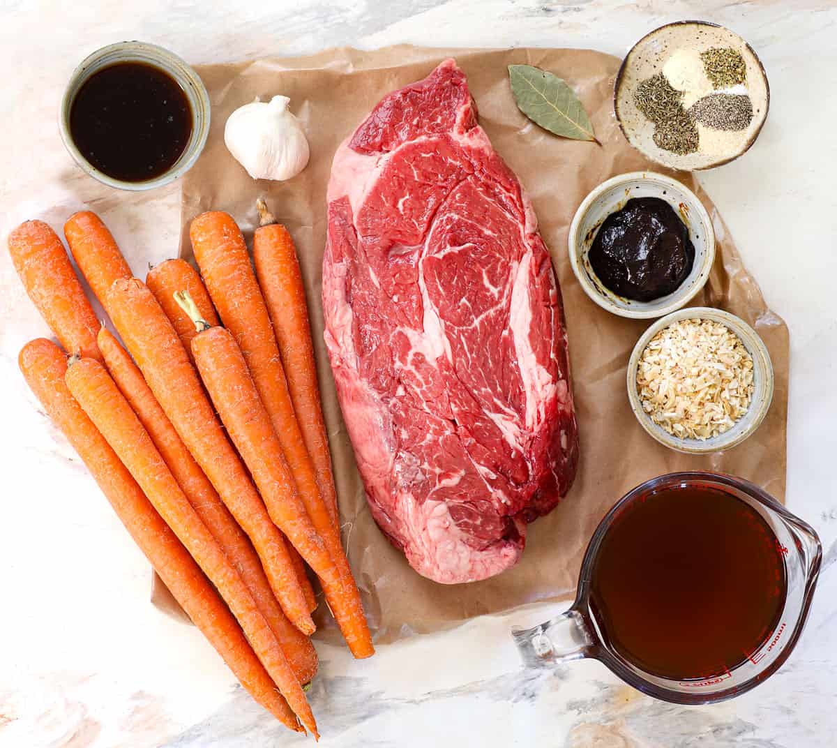 top view of pot roast ingredients: chuck roast, Worcestershire sauce, garlic, beef bouillon, dried minced onions, beef broth, spices, and carrots