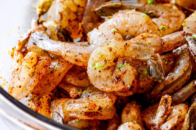 Cilantro Lime Shrimp Tacos EXPLODING with tangy, punchy flavor complimented by sweet refreshing Mango Salsa and silky Avocado Crema on your table in 35 minutes! You can prep the toppings ahead of time for a super quick and easy weeknight dinner that tastes better than any restaurant! showing how to make Shrimp Tacos by tossing raw shrimp with lime zest, garlic and seasonings in a glass bowl