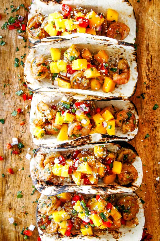 Cilantro Lime Shrimp Tacos EXPLODING with tangy, punchy flavor complimented by sweet refreshing Mango Salsa and silky Avocado Crema on your table in 35 minutes! You can prep the toppings ahead of time for a super quick and easy weeknight dinner that tastes better than any restaurant! top view of easy shrimp tacos recipe lined in a row on a brown cutting board