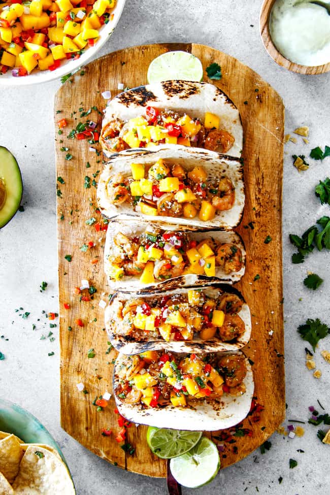 Cilantro Lime Shrimp Tacos EXPLODING with tangy, punchy flavor complimented by sweet refreshing Mango Salsa and silky Avocado Crema on your table in 35 minutes! You can prep the toppings ahead of time for a super quick and easy weeknight dinner that tastes better than any restaurant! easy Shrimp Tacos with Mango Salsa and Avocado lined in a row on a brown cutting board