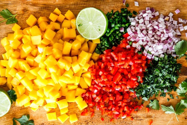 Cilantro Lime Shrimp Tacos EXPLODING with tangy, punchy flavor complimented by sweet refreshing Mango Salsa and silky Avocado Crema on your table in 35 minutes! You can prep the toppings ahead of time for a super quick and easy weeknight dinner that tastes better than any restaurant! showing how to make Shrimp Tacos by chopping mangos, red bell pepper, jalapeno, red onion, lime on a wood cutting board