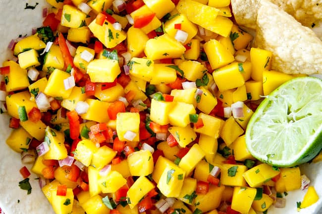 Cilantro Lime Shrimp Tacos EXPLODING with tangy, punchy flavor complimented by sweet refreshing Mango Salsa and silky Avocado Crema on your table in 35 minutes! You can prep the toppings ahead of time for a super quick and easy weeknight dinner that tastes better than any restaurant! showing how to make easy Shrimp Tacos by adding Mango Salsa to a white bowl