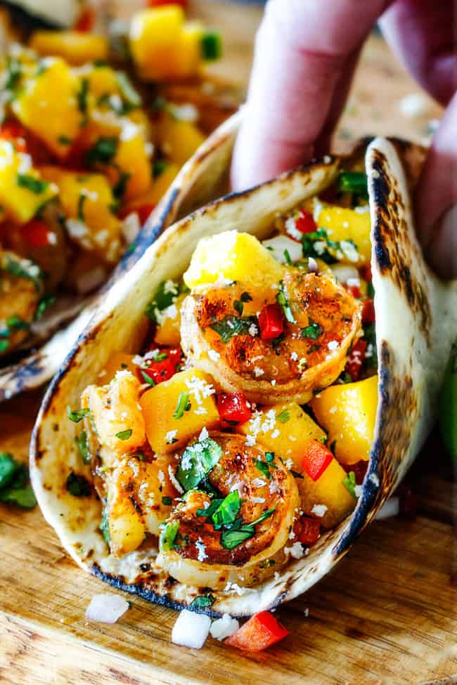 Cilantro Lime Shrimp Tacos EXPLODING with tangy, punchy flavor complimented by sweet refreshing Mango Salsa and silky Avocado Crema on your table in 35 minutes! You can prep the toppings ahead of time for a super quick and easy weeknight dinner that tastes better than any restaurant! up close of Shrimp Tacos with Mango Salsa and Avocado being grabbed by a hand to eat