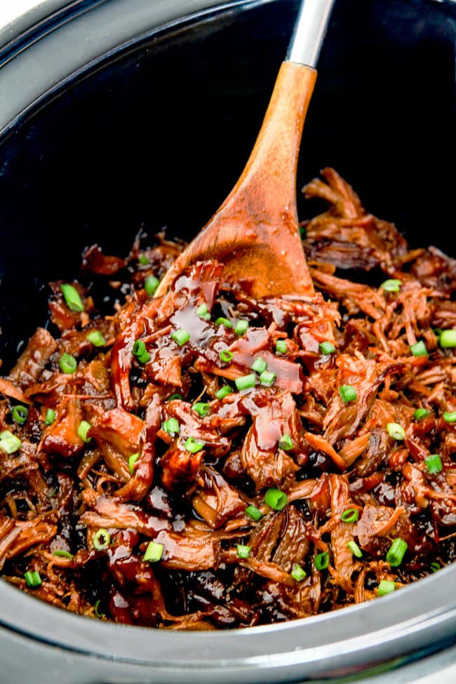 Slow cooker Pulled Pork in black slow cooker with a wooden spoon