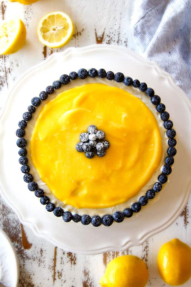top view of moist Lemon Blueberry Cake with white lemon cream cheese frosting and blueberries around the edges on a white cake stand
