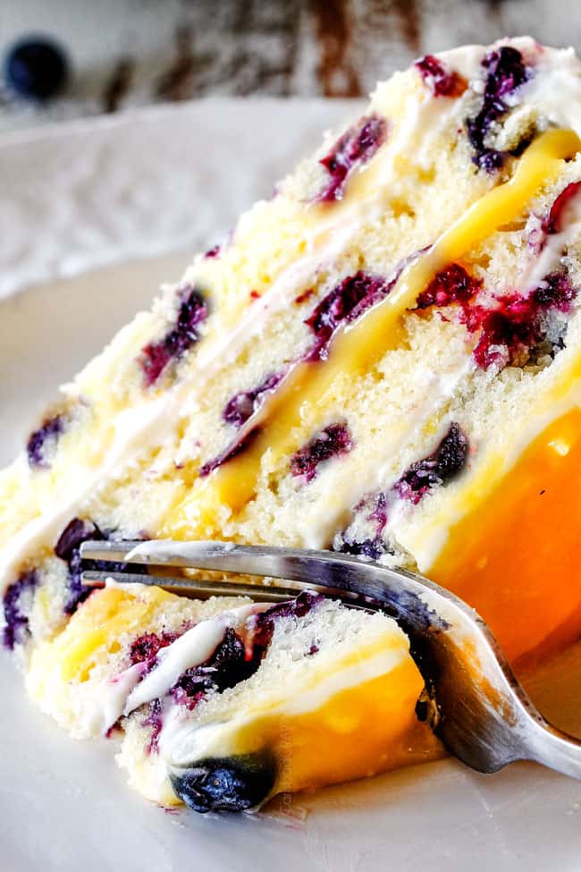 up close shot of slice of Lemon Blueberry Cake laying on its side on a white plate with a fork taking a bite