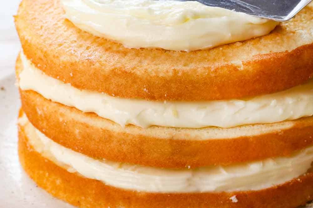 showing how to make coconut cake recipe by layering one cake then, topping with frosting, a second cake, then topping with a layer of frosting, then a third cake, and a layer of frosting