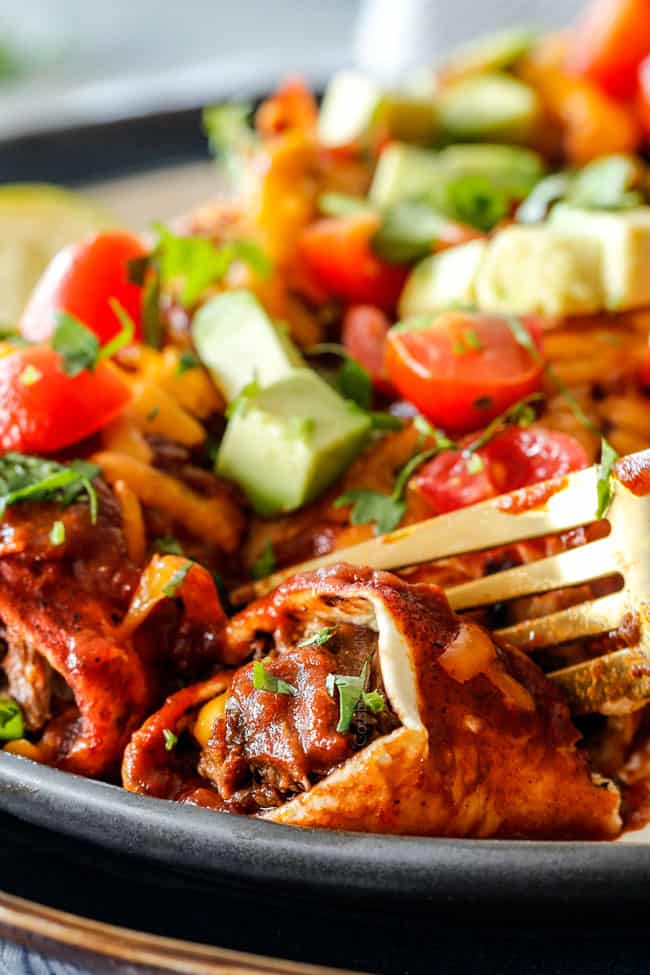 two best Beef Enchiladas on a white plate with a golden spoon garnished with tomatoes, avocado and cilantro