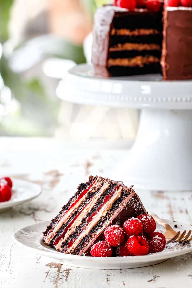 a far away shot of a a slice of moist Chocolate Raspberry Cake on its side showing layers of dark chocolate cake, raspberry jam filling, chocolate ganache and chocolate mousse with the cake on a white pedestal in the background