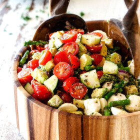 far away shot of Cucumber Tomato Salad recipe with cherry tomatoes, cucumbers, onion, asparagus and avocado in a wood bowl with wood tongs scooping salad
