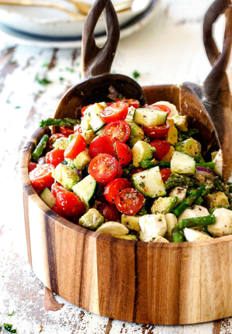 far away shot of Cucumber Tomato Salad recipe with cherry tomatoes, cucumbers, onion, asparagus and avocado in a wood bowl with wood tongs scooping salad