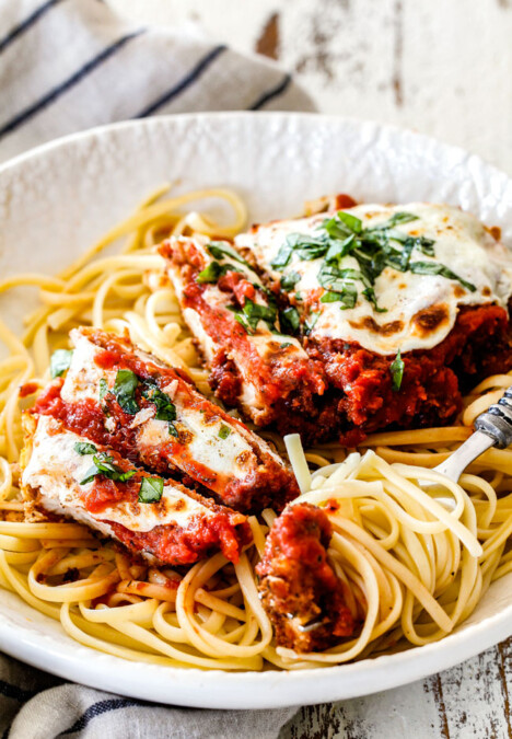 side view of Best chicken Parmesan partially sliced with spaghetti noodles in a white shallow bowl and a fork full of noodles