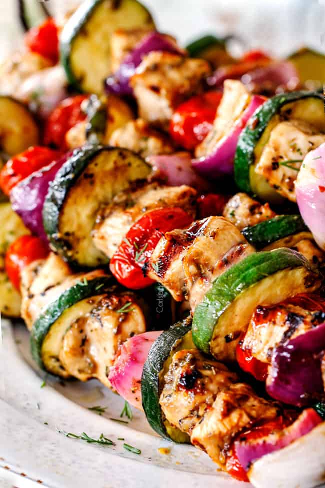 best chicken souvlaki skewers laying on a white plate with tomatoes, zucchini, tzatziki