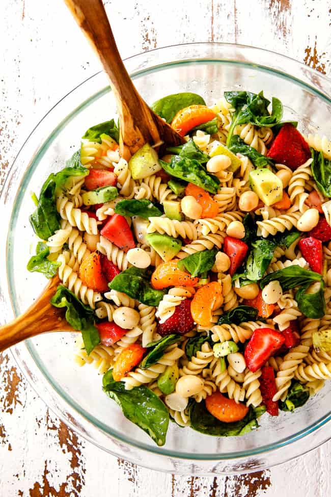 top view of pasta, strawberries, spinach, Mandarin oranges, avocados, macadamia nuts in a glass bowl with poppy seed dressing mixed together for pasta salad