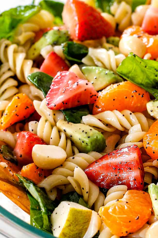 up close view of pasta, strawberries, spinach, Mandarin oranges, avocados, macadamia nuts in a glass bowl with poppy seed dressing mixed together for pasta salad