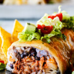 side view of wet burrito cut open with pulled pork, rice, black beans on a black plate garnished with lettuce, tomatoes, sour cream