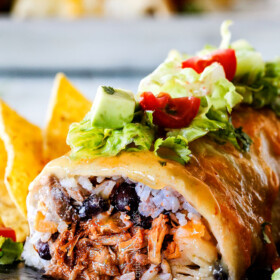 side view of wet burrito cut open with pulled pork, rice, black beans on a black plate garnished with lettuce, tomatoes, sour cream
