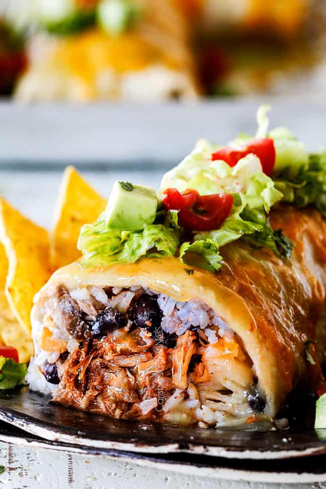 side view of wet burrito cut open with pulled pork, rice, black beans on a black plate garnished with lettuce, tomatoes, sour cream