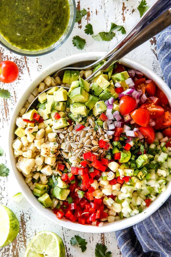 top view of corn salad ingredients in a white bowl with canned corn, avocados, tomatoes, jalapenos, cilantro, red onions, jalapeno, sesame seeds