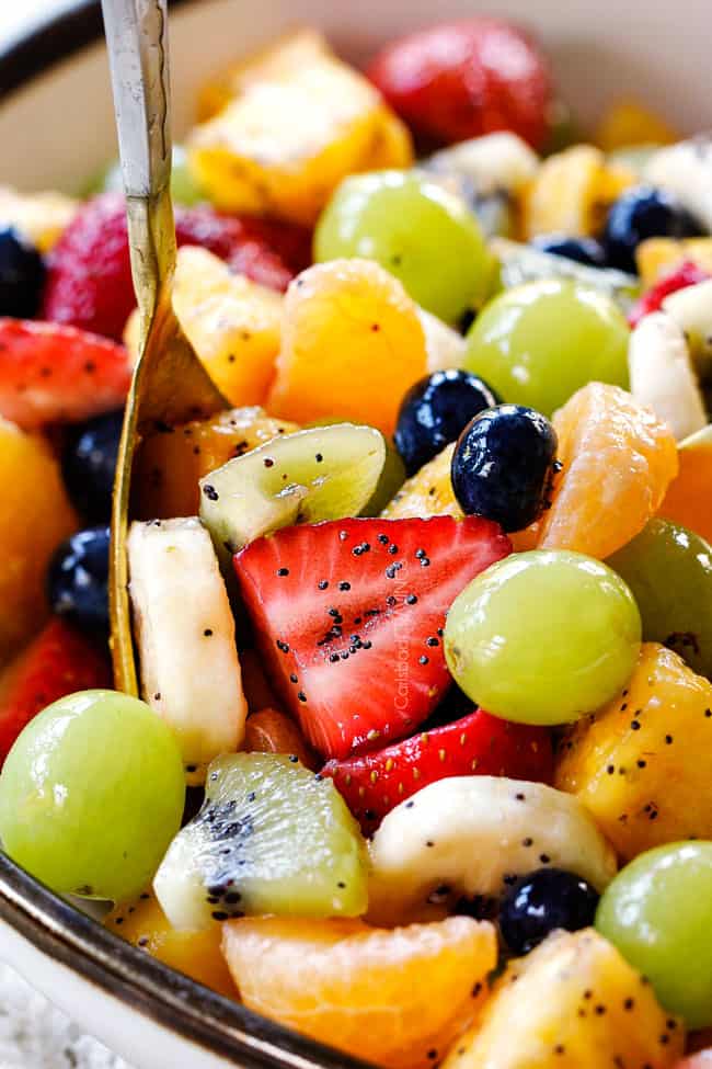 a gold spoon in a white bowl of easy fruit salad with pineapple, bananas, strawberries, blueberries, kiwis, oranges, pineapple