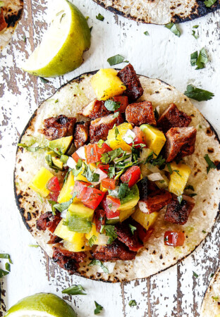 up close top view of al pastor pork on a corn tortilla with grilled pineapple, pico de gallo and avocado on a white board