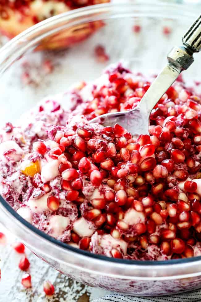 up close of a spoon scooping classic ambrosia salad with pineapple, mandarin oranges, mini marshmallows, cranberries and pomegranate seeds in a glass bowl