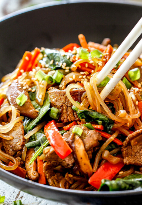 a bowl of Korean Spicy Noodles with beef, mushrooms, carrots, sesame seeds, green onions with white chopsticks