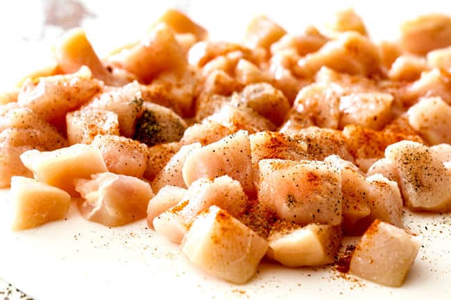 showing how to make basil chicken recipe by tossing chopped raw chicken with salt, pepper and paprika on a white cutting board