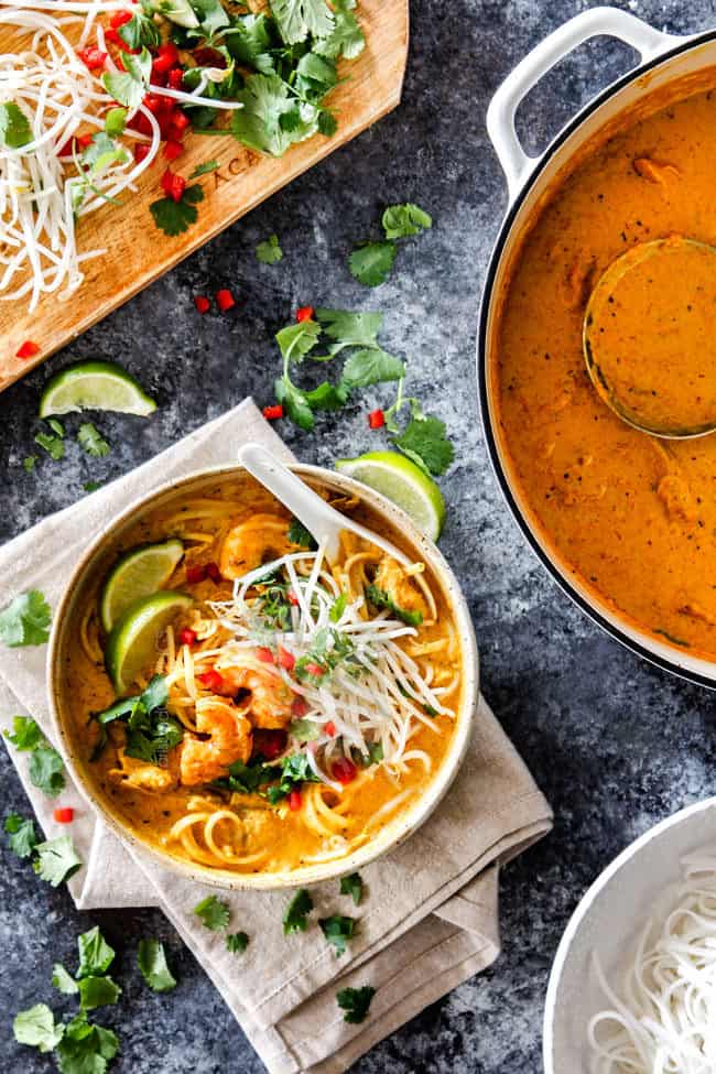 top view of a bowl of laksa soup with noodles, chicken and shrimp with a pot of laksa soup and a cutting board of bean sprouts, cilantro and peppers