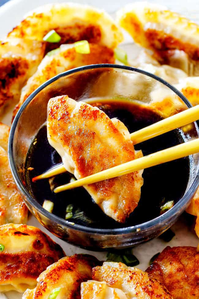 two chopsticks holding a potsticker getting ready to dunk it in potsticker dipping sauce on a tray with many potstickers