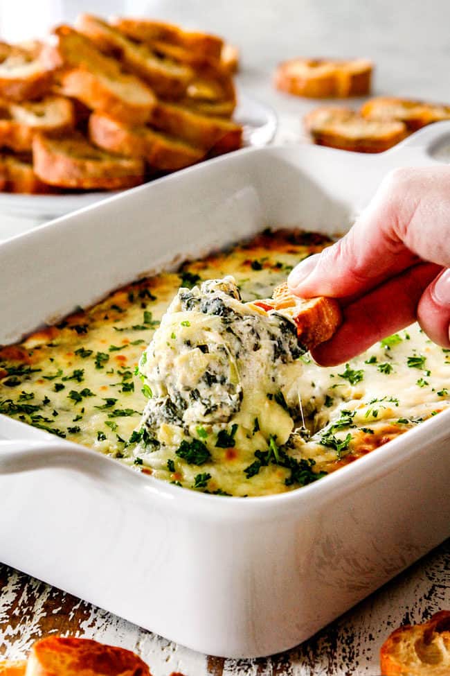 far away view of a hand picking up a piece of bread with cheesy artichoke dip on it