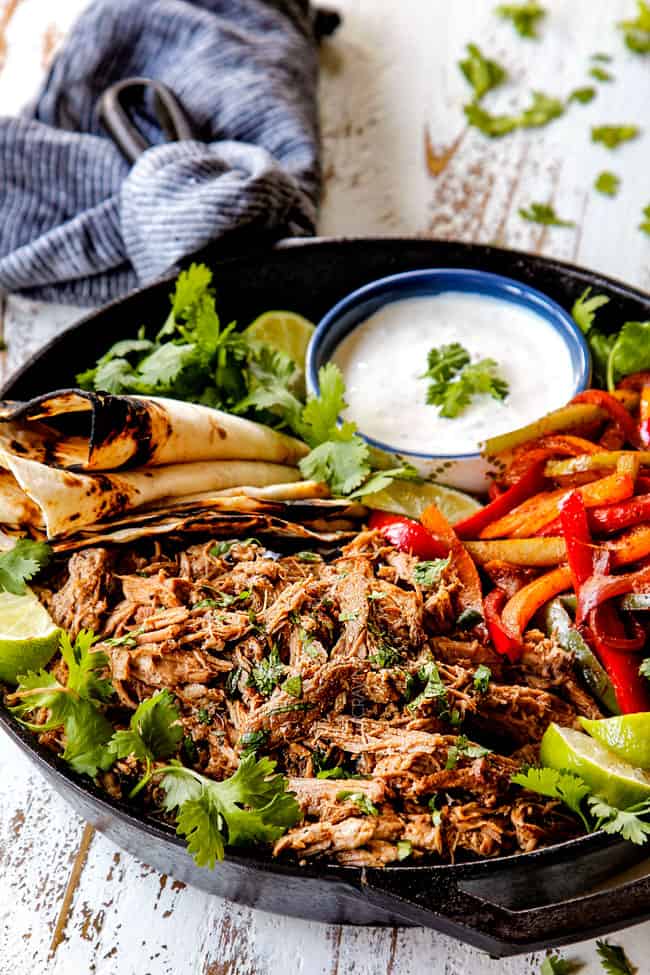 far away side view of pork fajitas in a skillet with salsa verde pork, bell peppers, onions, limes, cilantro and tortillas